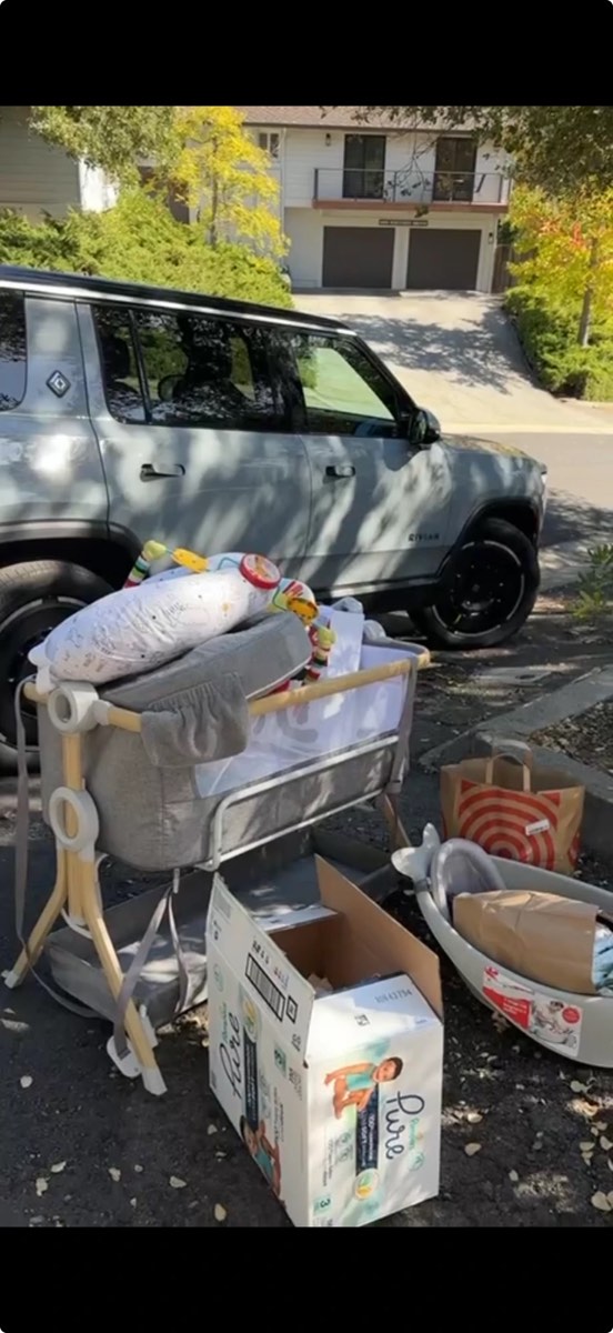 Baby things gathered by Merit neighbors for a pregnant mom — a bedside bassinet, baby bath, boxes of diapers and bags of gear next to a volunteer's car